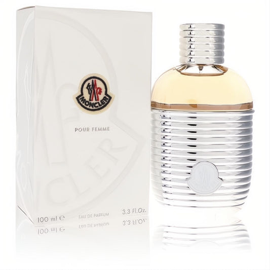 Luxurious Moncler Pour Femme Eau De Parfum, 100ml. Sleek silver ribbed bottle with warm amber liquid and iconic logo, beside its crisp white box.