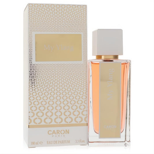 Luxurious Caron My Ylang 3.3 oz Eau De Parfum. Clear glass bottle with peachy fragrance, white cap, and gold details, beside its chic white and gold box.